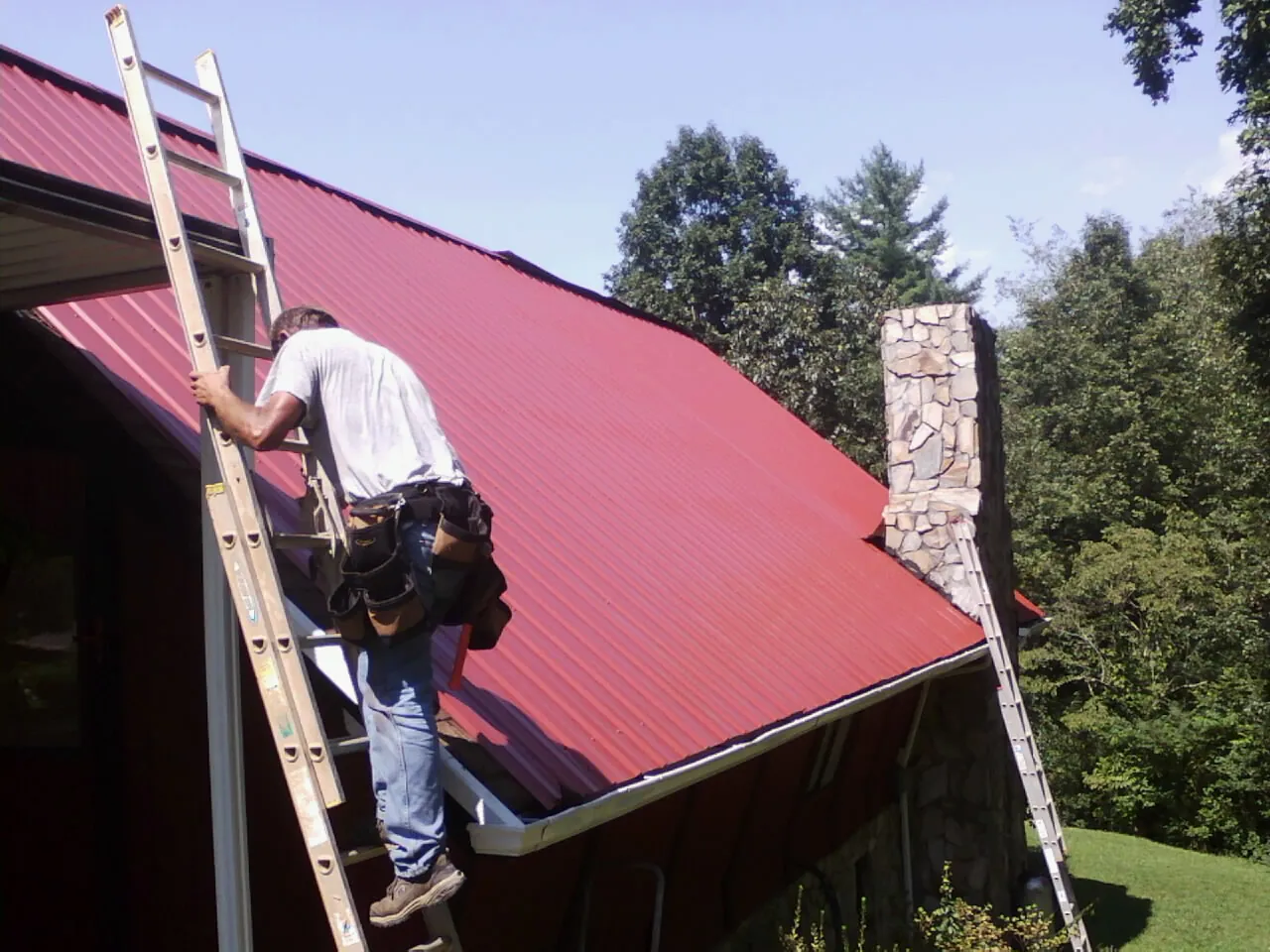 Roofer on a ladder beside a red metal roof and stone chimney — Soffit & Fascia Repair by Roofing Brothers in Ivoryton